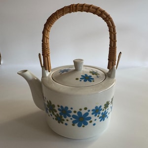 May include: A white ceramic teapot featuring a repeating pattern of blue and olive green flowers around the base and lid. The teapot has a curved spout and a tall, arched handle wrapped in light brown woven rattan material for a textured grip.