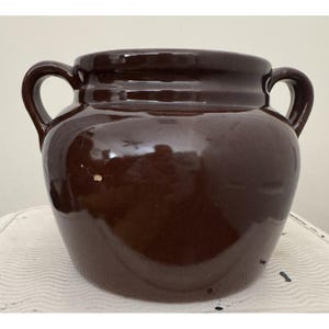 Może przedstawiać: Ciemnobrązowy, szkliwiony garnek ceramiczny o szerokim, zaokrąglonym korpusie i dwóch symetrycznych uchwytach bocznych. Naczynie ma gładką, odblaskową powierzchnię z grubym, wzmocnionym rantem przy górnym otworze, co nadaje mu solidny wygląd.