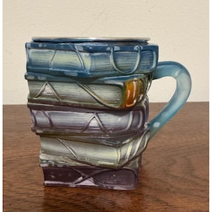 May include: A ceramic mug shaped like a stack of four books with a blue handle. The books are colored in shades of blue, purple, and grey with a metallic finish. The top book has a silver-colored rim, and the stack features embossed spine details.