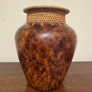 Könnte beinhalten: Eine Vase mit bauchigem Körper und einer gesprenkelten Oberfläche in Braun- und Dunkelbeigetönen. Der Hals der Vase ist mit hellbraunem, geflochtenem Rattan umwickelt, das mit dünnen, gleichmäßig verteilten vertikalen Rattanstreifen befestigt ist.