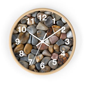 May include: Round wall clock with a light wood frame. The clock face is filled with smooth, colorful stones in shades of gray, brown, and tan. White numerals mark the hours, and white hands indicate the time.