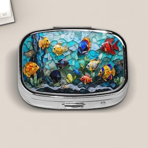 May include: A rectangular metal pill case with rounded corners featuring a vibrant mosaic-style print of colorful tropical fish swimming among aquatic plants and rocks in shades of blue, yellow, orange, and red against a light background.