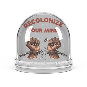 May include: A clear snow globe with a grey interior. It features two brown fists breaking chains, with the text "DECOLONIZE YOUR MIND" in red. The base is clear with a silver rim.