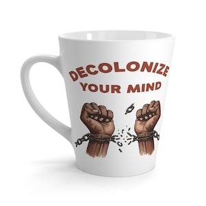 Könnte beinhalten: Ein weißer Keramik-Latte-Becher mit dem Text DECOLONIZE YOUR MIND in dunkelroten Großbuchstaben. Unter dem Text zeigt eine Illustration zwei dunkelhäutige Fäuste, die nach oben gereckt sind und jeweils aus dunkelgrauen Metallketten ausbrechen.