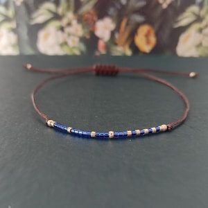 Könnte beinhalten: Minimalistisches Armband mit dunkelblauen und goldenen Perlen auf einer braunen Kordel. Das verstellbare Armband hat einen Schiebeknotenverschluss. Die Perlen sind gleichmäßig auf der Kordel verteilt. Schlichtes, elegantes Schmuckstück.