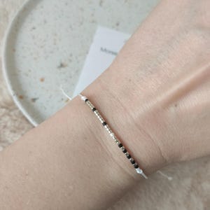 Könnte beinhalten: Ein minimalistisches Armband mit kleinen schwarzen, silbernen und weißen Perlen auf einer dünnen weißen Schnur. Das Armband wird an einem Handgelenk gezeigt und unterstreicht sein schlichtes Design und seine Farbgebung.