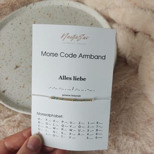 Könnte beinhalten: Eine weiße Karte mit dem Text "NastjaSar HANDMADE JEWELRY Morse Code Armband Alles liebe geheime Botschaft" wird vor einem weißen Schnurarmband mit goldenen und türkisfarbenen Perlen gehalten. Die Karte zeigt auch das Morsealphabet.