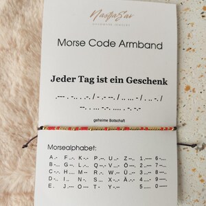 Könnte beinhalten: Ein Morsecode-Armband mit roten und goldenen Perlen wird auf einer Karte gezeigt. Auf der Karte steht "Morse Code Armband" und "Jeder Tag ist ein Geschenk" auf Deutsch. Das Morsealphabet ist ebenfalls aufgeführt.