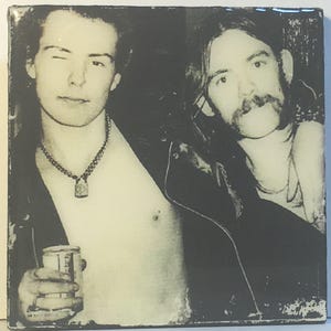 May include: Black and white photo of two men wearing leather jackets. The man on the left is holding a can of beer. The man on the right has a mustache and is wearing a necklace.