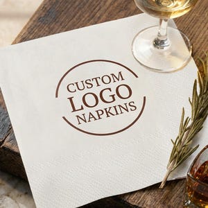 May include: White square napkin with the text "CUSTOM LOGO NAPKINS" in a brown circular design. A glass of white wine, a shot of amber liquor, and a sprig of rosemary are on a wooden surface.