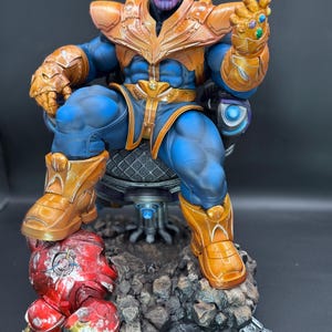 May include: A detailed Thanos action figure sitting on a dark throne, wearing blue and gold armor with a gauntlet featuring colorful stones. The figure rests on a rocky base with a damaged red and gold helmet piece lying at the feet of the character.