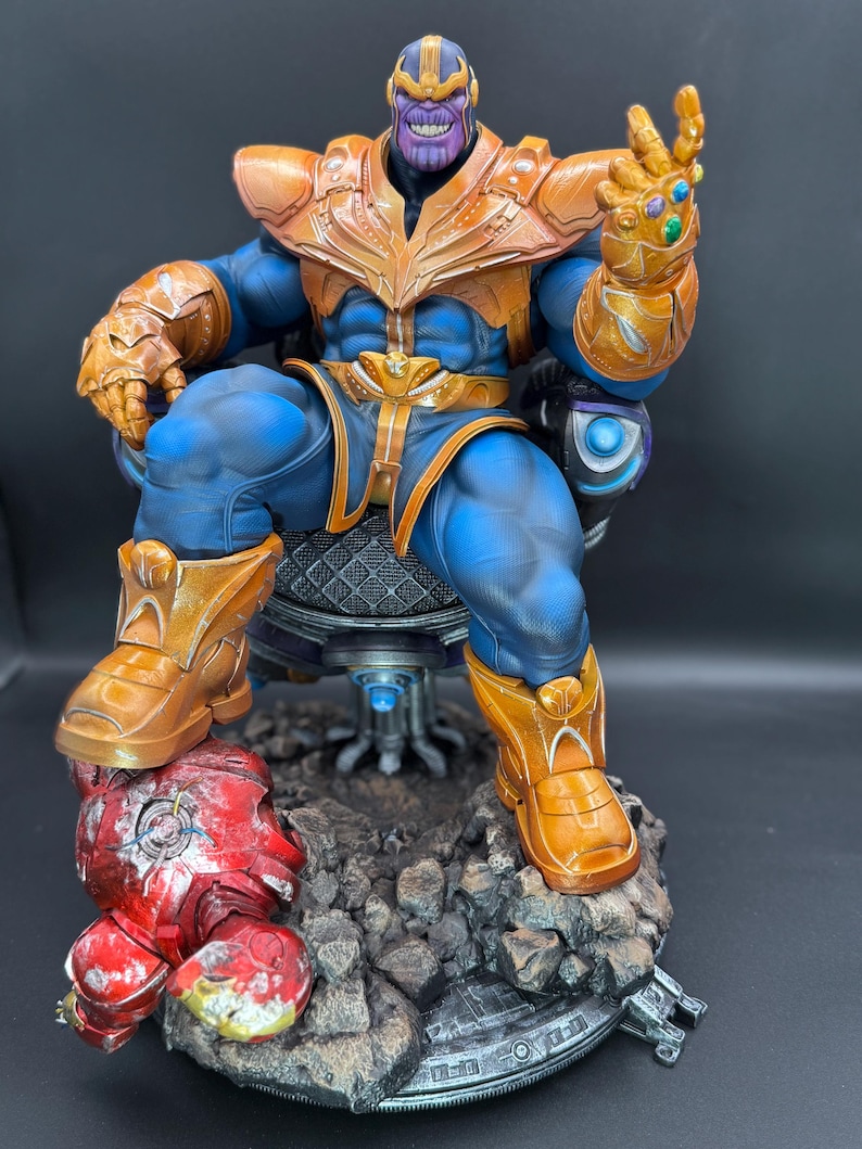 May include: A detailed Thanos action figure sitting on a dark throne, wearing blue and gold armor with a gauntlet featuring colorful stones. The figure rests on a rocky base with a damaged red and gold helmet piece lying at the feet of the character.