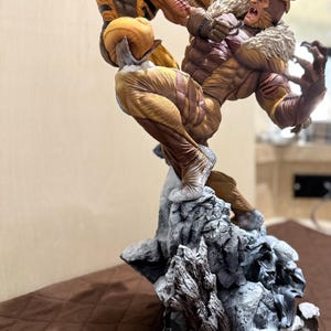 May include: A dynamic action statue featuring Wolverine in a yellow and brown suit with extended claws leaping toward Sabretooth, who wears a brown suit with a fur collar. They are mounted on a detailed rocky base with a circular red and yellow X-Men logo.