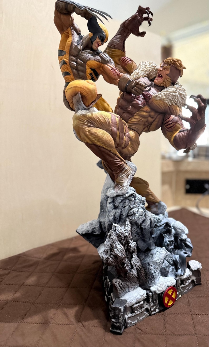 May include: A dynamic action statue featuring Wolverine in a yellow and brown suit with extended claws leaping toward Sabretooth, who wears a brown suit with a fur collar. They are mounted on a detailed rocky base with a circular red and yellow X-Men logo.