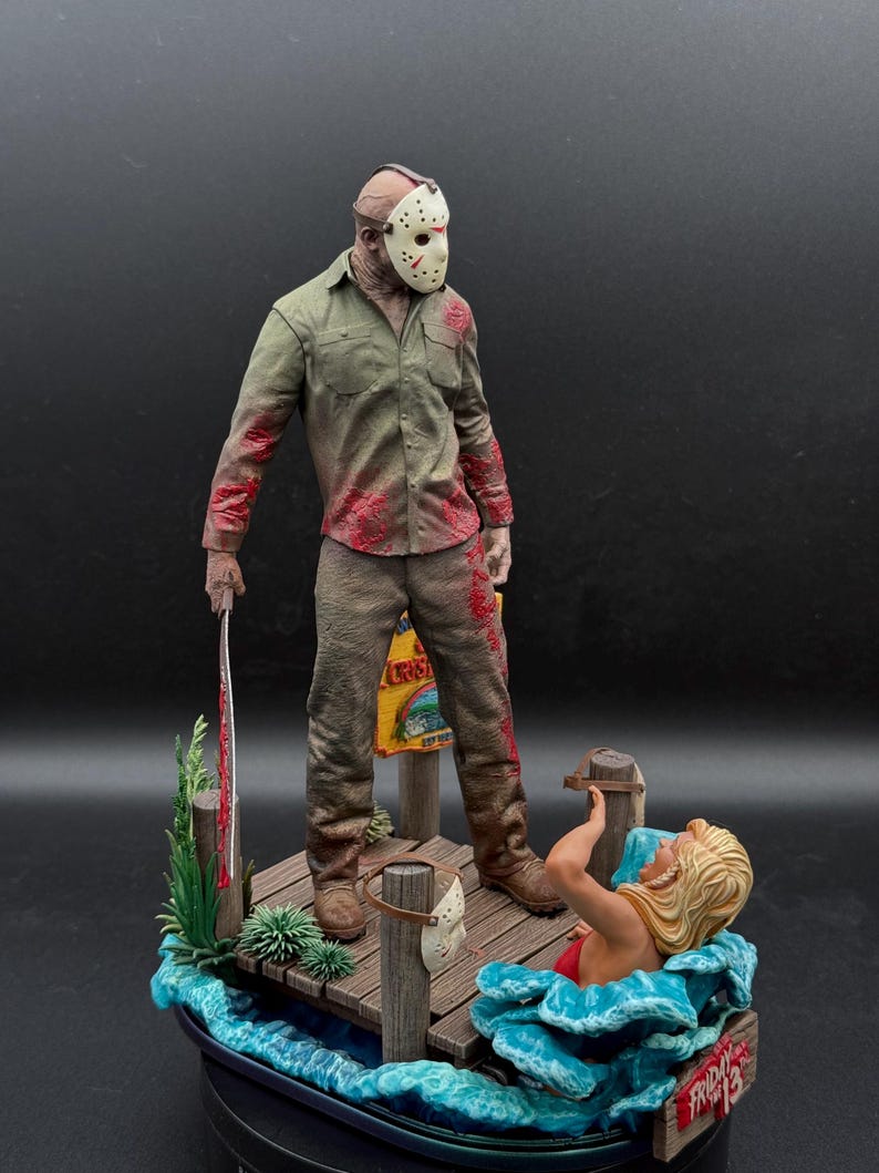 May include: A detailed figurine set featuring a character wearing a white hockey mask and a blood-stained olive green shirt and brown pants, holding a machete on a wooden dock. A blonde figure is in the blue water, with a Friday the 13th sign.