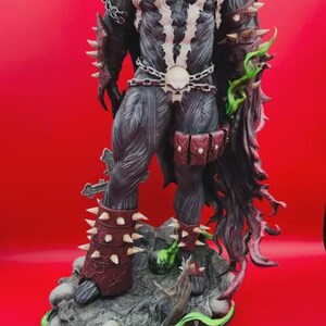 May include: A Spawn action figure standing on a rocky base with skulls and green energy effects. The figure features a black suit with white chest markings, a red cape, spiked arm guards, a skull belt with chains, and a pouch on the left thigh.