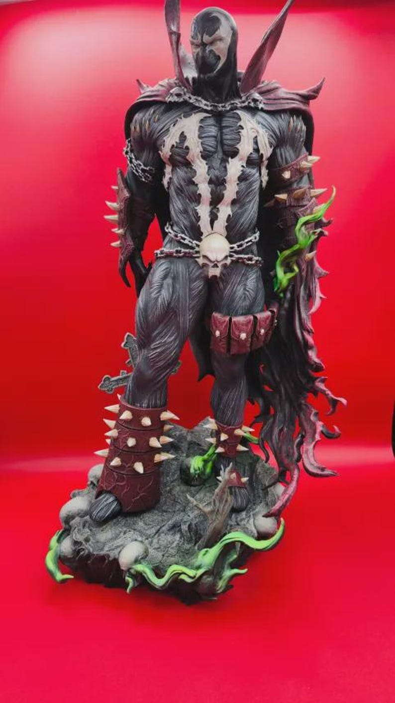 May include: A Spawn action figure standing on a rocky base with skulls and green energy effects. The figure features a black suit with white chest markings, a red cape, spiked arm guards, a skull belt with chains, and a pouch on the left thigh.