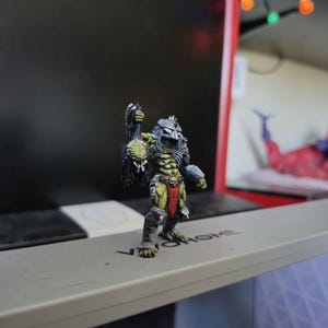 May include: A miniature figurine of a green-skinned creature with a metallic helmet, dark armor, and a red loincloth. The figure holds a dark, textured object in its raised right hand and stands on a gray surface with the text VIVOHOME printed on it.