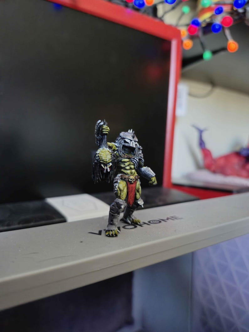 May include: A miniature figurine of a green-skinned creature with a metallic helmet, dark armor, and a red loincloth. The figure holds a dark, textured object in its raised right hand and stands on a gray surface with the text VIVOHOME printed on it.