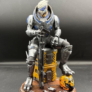May include: A detailed figurine of a humanoid alien character with grey skin and blue facial markings, wearing metallic silver and blue armor. The figure sits on a yellow industrial crate, holding a long rifle, with a yellow helmet and shell casings.