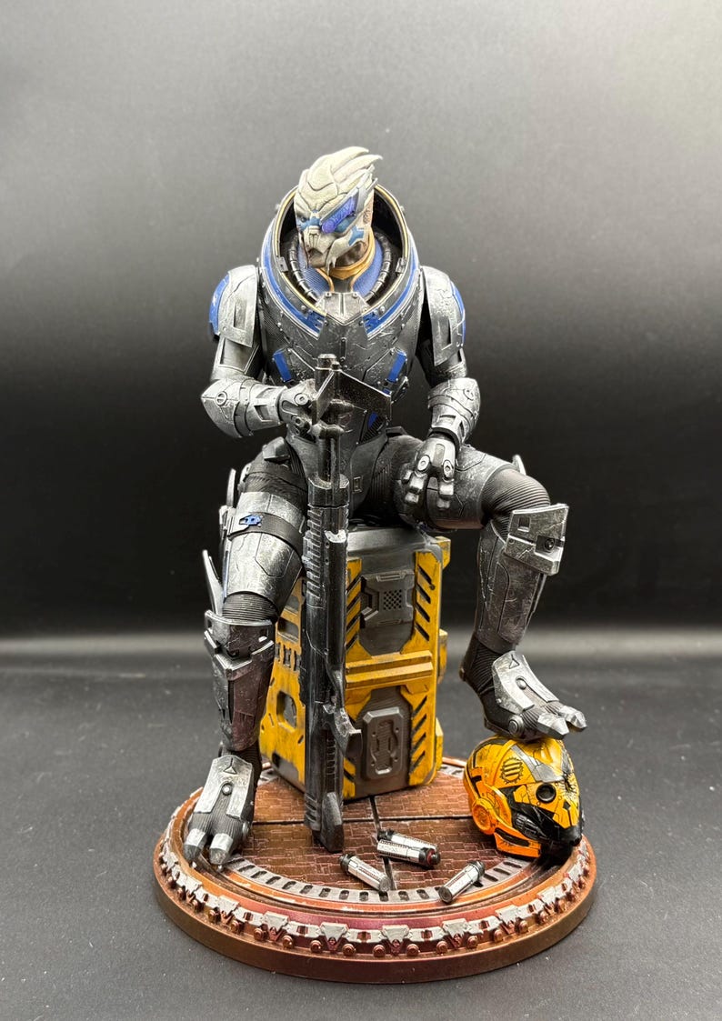 May include: A detailed figurine of a humanoid alien character with grey skin and blue facial markings, wearing metallic silver and blue armor. The figure sits on a yellow industrial crate, holding a long rifle, with a yellow helmet and shell casings.