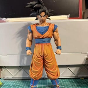 May include: A standing anime character figurine featuring spiky black hair, wearing an orange gi with a blue undershirt, blue wristbands, a blue sash, and blue boots with red accents. The figure has defined muscular arms and a serious facial expression.