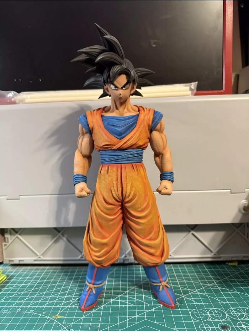 May include: A standing anime character figurine featuring spiky black hair, wearing an orange gi with a blue undershirt, blue wristbands, a blue sash, and blue boots with red accents. The figure has defined muscular arms and a serious facial expression.