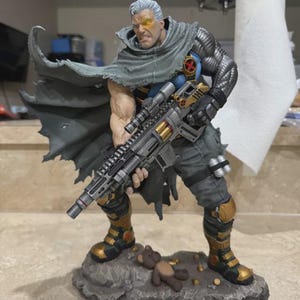 May include: A detailed figurine of a muscular character with silver hair, a glowing yellow eye, and a metallic left arm. The figure wears a tattered gray cape, blue shirt with an X-logo, and holds a large rifle. It stands on a rocky base with a teddy bear.