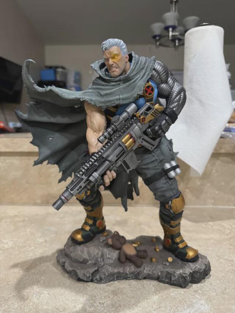 May include: A detailed figurine of a muscular character with silver hair, a glowing yellow eye, and a metallic left arm. The figure wears a tattered gray cape, blue shirt with an X-logo, and holds a large rifle. It stands on a rocky base with a teddy bear.