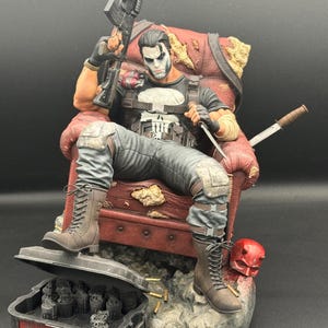 May include: A detailed figurine of The Punisher sitting in a distressed red armchair, holding a rifle and a knife. The character wears a black shirt with a white skull emblem, grey tactical pants, and brown boots. A red mask and a case of grenades sit nearby.