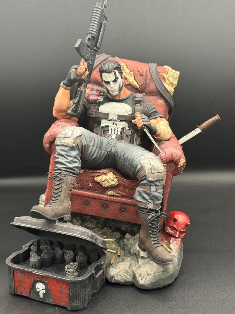 May include: A detailed figurine of The Punisher sitting in a distressed red armchair, holding a rifle and a knife. The character wears a black shirt with a white skull emblem, grey tactical pants, and brown boots. A red mask and a case of grenades sit nearby.