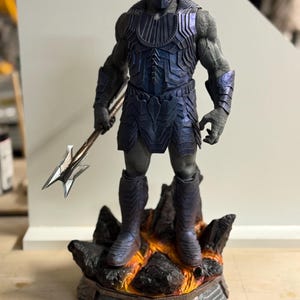 May include: A gray-skinned humanoid figure with glowing red eyes, wearing dark blue metallic armor and holding a silver spear. The figure stands on a rocky base featuring orange lava-like details, with a textured, segmented design on the armor.