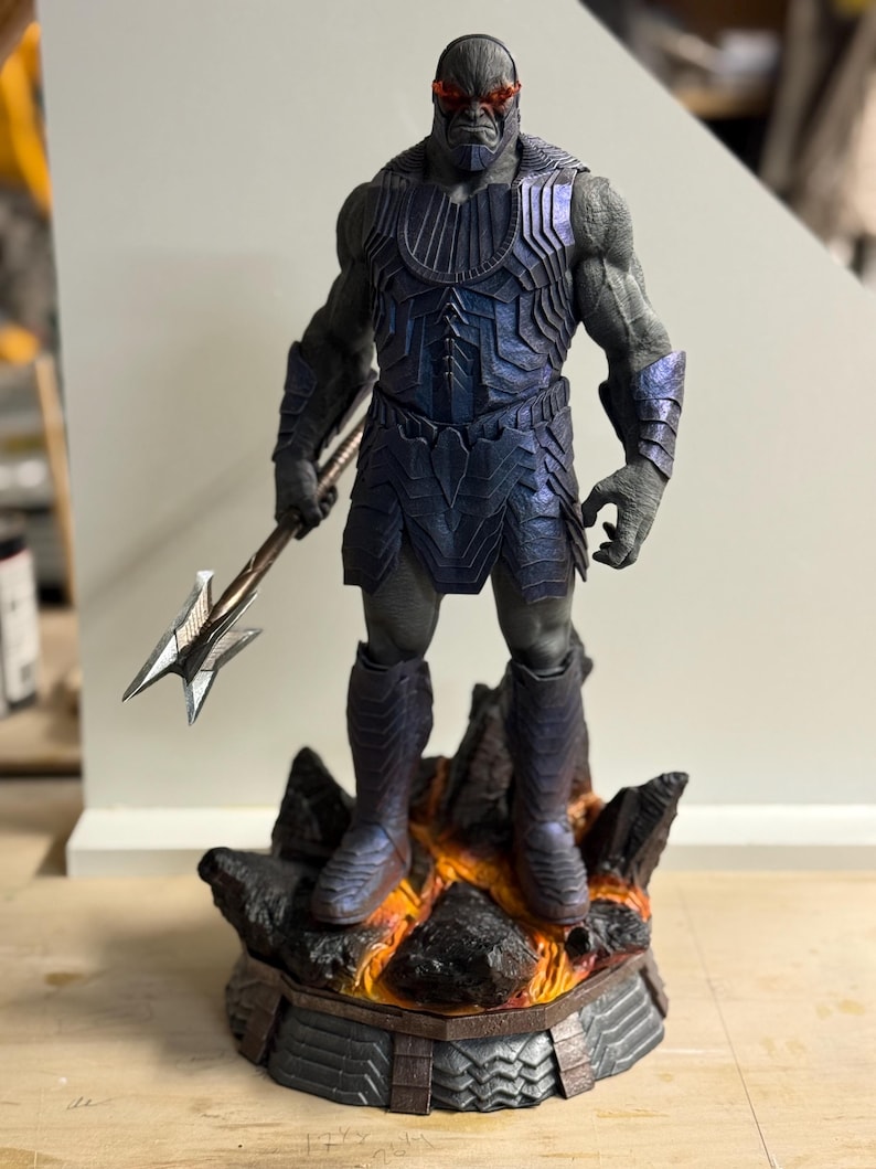 May include: A gray-skinned humanoid figure with glowing red eyes, wearing dark blue metallic armor and holding a silver spear. The figure stands on a rocky base featuring orange lava-like details, with a textured, segmented design on the armor.