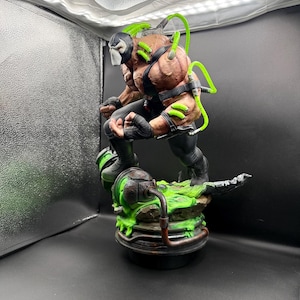May include: A highly detailed Bane action figure statue featuring a muscular physique, a black mask, and black tactical gear. Bright neon green tubes connect to the back and arms, while the figure stands on a base with green liquid effects.