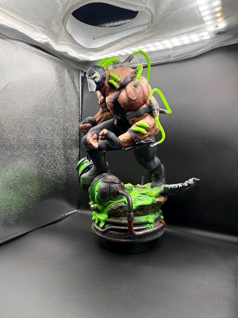 May include: A highly detailed Bane action figure statue featuring a muscular physique, a black mask, and black tactical gear. Bright neon green tubes connect to the back and arms, while the figure stands on a base with green liquid effects.