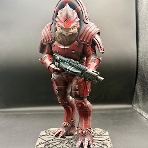 May include: A detailed figurine of a reptilian humanoid warrior wearing weathered red armor and a large back plate. The figure holds a dark green rifle with both hands and stands on a textured, rectangular stone-like base with carved patterns.
