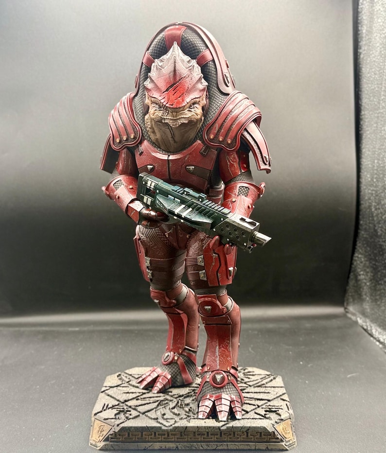 May include: A detailed figurine of a reptilian humanoid warrior wearing weathered red armor and a large back plate. The figure holds a dark green rifle with both hands and stands on a textured, rectangular stone-like base with carved patterns.