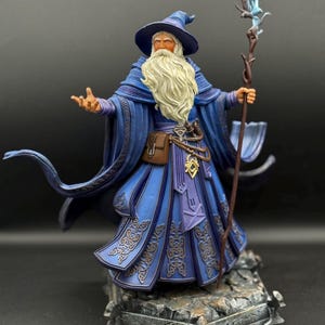 May include: A detailed figurine of a wizard with a long white beard, wearing a blue robe and pointed hat. The figure holds a wooden staff topped with a glowing blue crystal and stands on a rocky base, featuring intricate knotwork patterns.