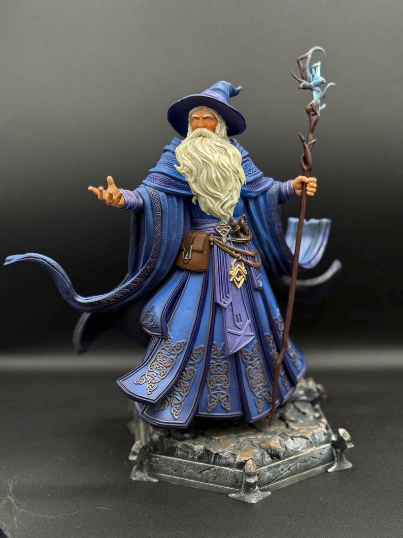 May include: A detailed figurine of a wizard with a long white beard, wearing a blue robe and pointed hat. The figure holds a wooden staff topped with a glowing blue crystal and stands on a rocky base, featuring intricate knotwork patterns.