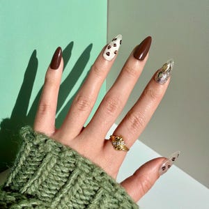 Puede incluir: Una mano con esmalte de uñas marrón y blanco, un anillo de oro con piedras transparentes y un esmalte de uñas con purpurina plateado. La mano lleva un suéter de punto verde.