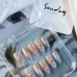 Puede incluir: Una caja de plástico transparente con diez uñas postizas iridiscentes, nacaradas y blancas con pedrería y adornos en 3D. La caja tiene el texto "Sunday Studio" en la tapa.