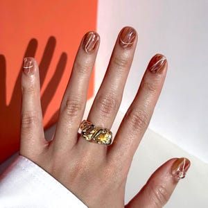 Puede incluir: Una mano con esmalte de uñas marrón y blanco con un anillo de oro en el dedo medio.