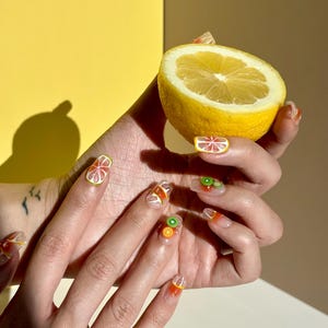 May include: A hand with clear nail polish and fruit-themed nail art is holding a lemon wedge. The nails are decorated with orange, kiwi, and lemon slices.