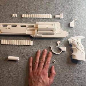 Pode incluir: Um revólver de brinquedo branco desmontado, com vários componentes dispostos. As peças incluem o corpo principal, uma empunhadura com design de asa, trilhos e outras peças pequenas. Os componentes estão dispostos sobre uma superfície cinza.