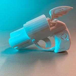 Op de afbeelding: Een witte 3D-geprinte handpistool met een gevleugelde figuur en een hartvormig detail. Het pistool heeft een cilindrische loop en een trekkerbeugel. De achtergrond is een verloop van blauw en oranje.
