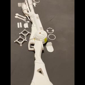 May include: A disassembled white toy rifle with various components, including a scope, barrel, and stock. The parts are arranged on a dark surface, suggesting a model kit or a project in progress. The design features a futuristic aesthetic.