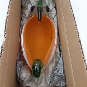 May include: A decorative glass bird, orange and clear with a green tail and beak, and blue eyes. The object is in a cardboard box with protective wrapping.