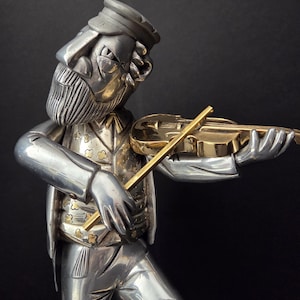 May include: A silver-toned figurine of a musician playing a violin. The figure wears a hat, vest with gold accents, and pants. The violin is gold-colored, and the bow is held in the right hand. The background is black.