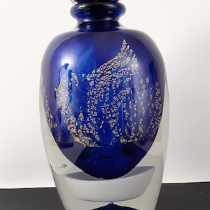 May include: A decorative glass bottle with a dark blue body and a clear base. The blue section features gold flecks and a matching spherical stopper. The bottle has a unique, artistic design.
