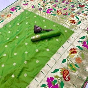 May include: A vibrant green silk saree with a gold border and floral embroidery. The saree features a detailed design of colorful flowers and leaves. A rolled-up section of the saree and a decorative object are also visible.
